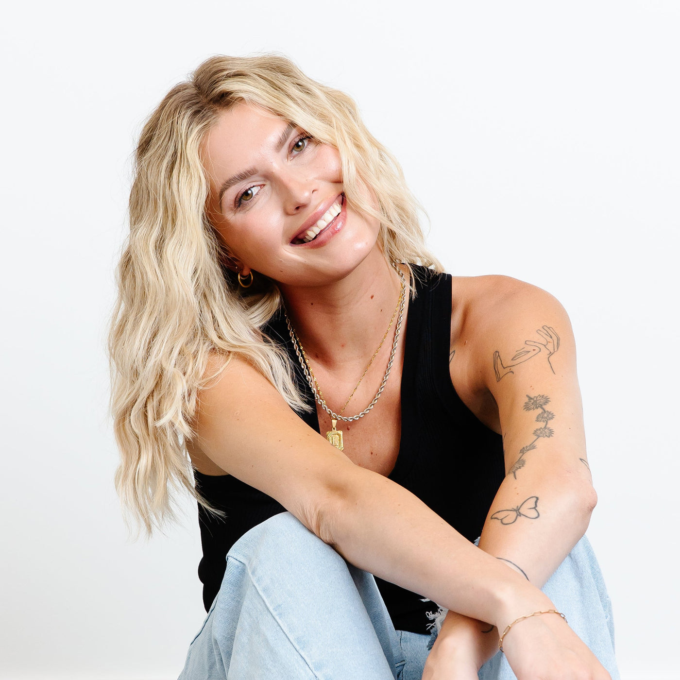 A woman with tattoos, dressed in a black t-shirt and jeans, sits gracefully on a white floor, with wavy blonde hair.