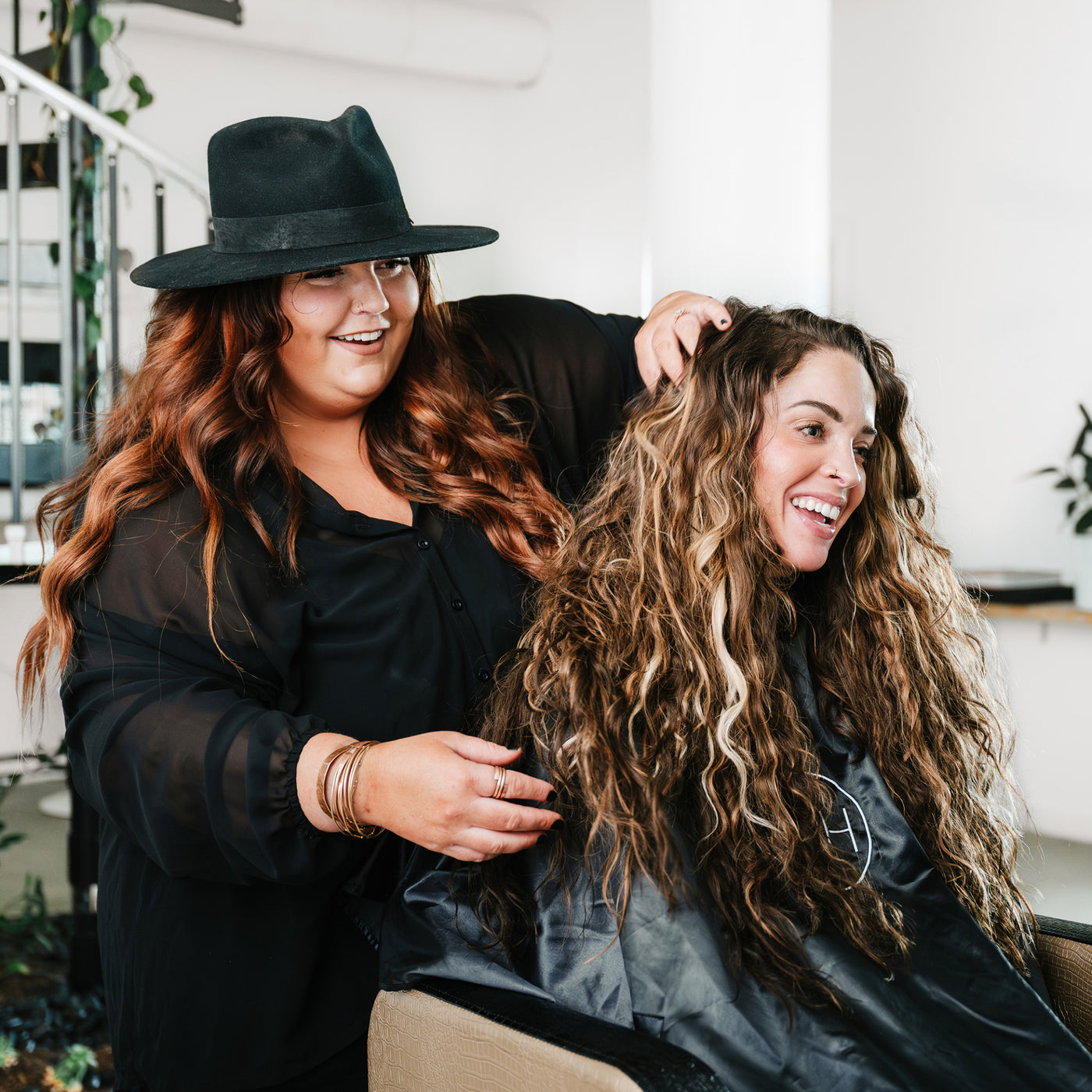 A hairstylist in a black hat and outfit styling a client’s Kera-Link hair extensions in a modern salon setting.