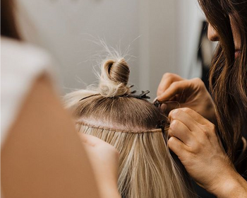 A hairstylist applying hair extensions to another woman's hair in a salon.